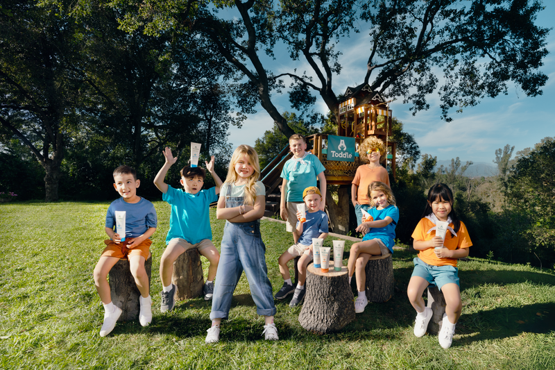 Group of kids gathered outdoors in front of a Toddle-branded treehouse holding the full Toddle kids skincare lineup — sunscreen, lotion, and 3-in-1 wash.